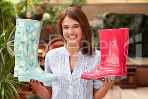 Ive just gotta have both pairs. A smiling woman holding up different gumboots while standing in a plant nursery.