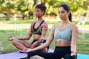 Focusing on mindfulness and the breath. Cropped shot of two attractive young women sitting next to each other and meditating in the park.