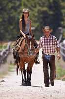 Riding 101. An attractive young cowgirl riding a horse on a ranch while a cowboy walks alongside.