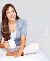 Cute relaxed female drinking coffee and looking away in thought. Happy thoughtful woman sitting legs crossed on bed and drinking a cup of coffee.