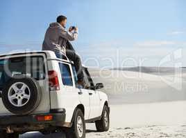 Capturing the scenery. Cropped shot of a young man sitting on his car roof while on a roadtrip.