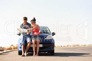 Finding their way to that perfect destination. A man and a woman parked on the side of the road and reading a map together while seated on the bonnet of their car.