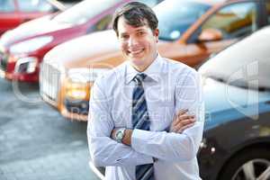 He can give you a great car deal. A man smiling confidently while standing in the lot of his dealership.