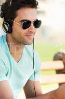 Tuning out to his favorite tunes. A smiling young man wearing sunglasses sitting on a bench in a park and listening to music through his headphones.