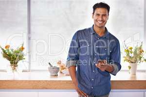 Maybe I should invite some friends over for dinner. Portrait of a handsome holding a mobile phone while standing in the kitchen.