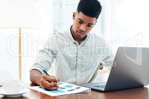 Every work task gets his full focus. Shot of a young businessman using a laptop and going over paperwork in a modern office.