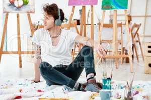 Getting some artistic inspiration through music. Full length shot of a handsome young artist sitting alone and looking contemplative while listening to music through headphones.