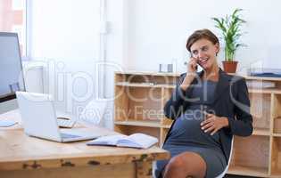 The baby is really kicky today. A pregnant businesswoman having an enjoyable conversation on the phone at her office desk.