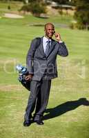 Doing business on the course. Shot of an african wearing a suit and talking on the phone while carrying a golf bag on a golf course.