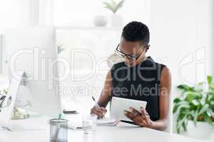 Stay organised, stay in control. Shot of a confident young businesswoman writing in a notebook and using a digital tablet in a modern office.