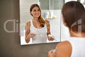 Step one to a brilliant smile. Over the shoulder portrait of an attractive young woman brushing her teeth in a bathroom mirror.