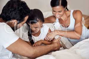 Time for some silly fun. Shot of parents tickling their little daughter while lying in bed together at home.