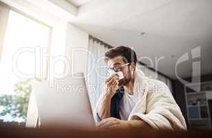 The flu has got him working from home today. Shot of a sickly young businessman blowing his nose with a tissue while working from home.