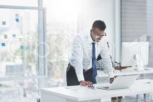 Theres always reward in patience and hard work. Shot of a young businessman standing over his office desk and looking at figures on a laptop.