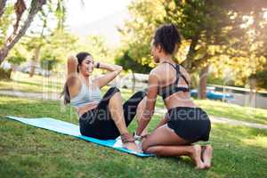 Keeping defined abs in mind. Full length shot of two attractive young women sitting and performing core exercises while in the park during the day.