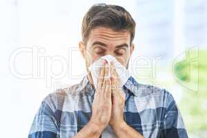 Going t have to stock up on tissues. Portrait of an uncomfortable looking young man blowing his nose with a tissue inside of a building during the day.