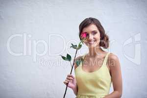 Shes a flower type of girl. Studio shot of a cheerful young woman holding a pink rose next to her face while standing against a grey background.