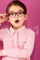 Shes seriously surprised. A little girl with expression of surprise holding the edge of her spectacles against a pink background.