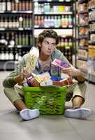 Which one do I need more. A young man sitting on the floor anxiously overwhelmed by grocery shopping.