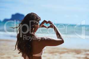 I just love the beach. Shot of a young woman shaping a heart while looking at the scenery at the beach.