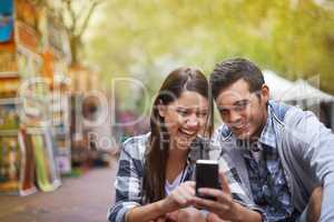 Lets capture this moment. A happy young couple sitting outside looking at pictures on their cellphone.