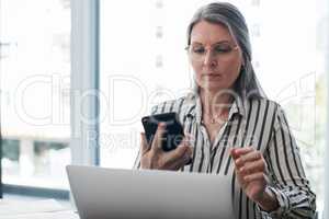 I got money to validate, icy like carrot cake. Shot of a mature businesswoman using her smartphone and laptop in a modern office.