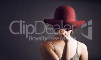 Want me to reveal the mystery. Studio shot of an unrecognizable young woman covering her face with a hat against a grey background.