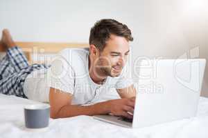 Coffee and wifi, all he needs to stay woke. Shot of a young man using a laptop while having coffee on his bed at home.