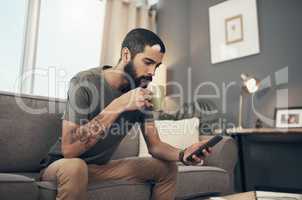 Connect to all things current with the ease of technology. Shot of a young man having coffee and using a smartphone on the sofa at home.