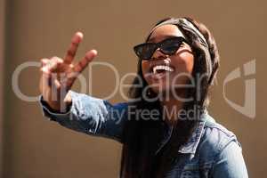 Be bold, be brave, be you. Cropped portrait of an attractive young woman wearing sunglasses and making a peace sign while standing against a wall alone.