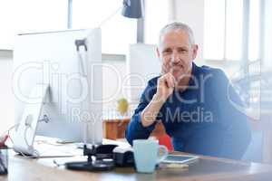 Leading a creative campaign into financial success. Mature creative professional looking positively at the camera while sitting at his desk in a bright work space.