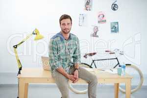 Portrait of a casually-dressed young man sitting on his desk in an office. The commercial designs displayed in this image represent a simulation of a real product and have been changed or altered enough by our team of retouching and design specialists so 