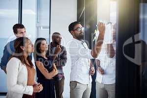 Hes showing them a new way of doing things. Shot of a young businessman giving a demonstration on a glass wall to his colleagues in a modern office.