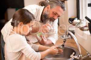 Washing the germs away. Shot of a girl washing her hands in the kitchen sink as her grandfather stands by.