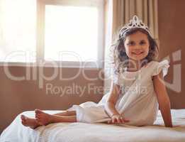 Is she not just the cutest princess youve ever seen. Portrait of an adorable little girl wearing a tiara and sitting on a bed at home.