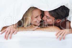 Keeping cosy. A couple lying under a duvet cover and cuddling affectionately while looking into each others eyes.