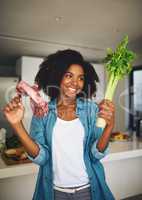 We have all the ingredients well ever need. Shot of a cheerful young woman holding celery in one hand and a piece of meat in the other while standing in the kitchen at home.