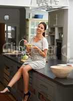 Feeling healthy and happy. Portrait of a young woman sitting on the kitchen countertop and eating a bowl of salad.