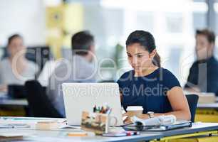 Immersed in her work. Shot of a designer sitting at her desk working on a laptop with colleagues in the background.