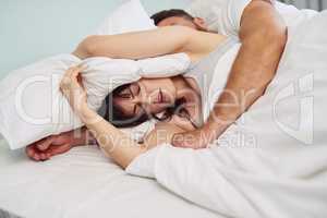 I just want to sleep in peace. Shot of a uncomfortable looking young woman putting a pillow over her head to try and sleep while her partner snores in bed.