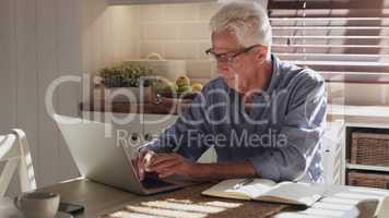I better get this to the client quick. Shot of a senior man sitting alone in the kitchen and using his laptop to work from home.