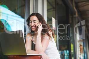 Booking her accommodation in a foreign city. Shot of a young woman speaking on her cellphone while using her laptop outside a coffee shop.