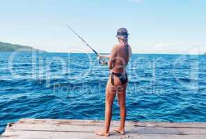 Shes what you call the ultimate catch. Rearview shot of an attractive young woman standing on a pier and fishing near an ocean.