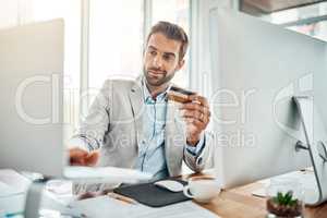 The ease of online banking. Shot of a handsome young businessman using a computer while holding a credit card in an office.