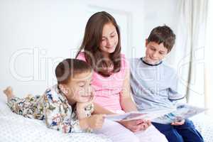 Sharing their interests together. Three siblings reading a magazine together happily in bed.