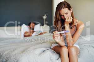 Please be negative.... Shot of a woman looking worried while holding a pregnancy test with her partner lying in the background.