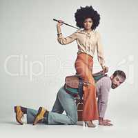 Get moving. A studio shot of an attractive woman in 70s wear riding a handsome man wearing a saddle while using a riding crop.