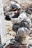 Trained in combat. A soldier crouched down next to a broken down wall and pointing his gun into the distance.
