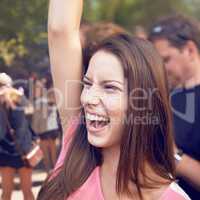 Whoohoo. A beautiful young woman smiling and enjoying music at a festival with arm raised in the air and crowd in the background.
