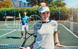 I'm always on the winning side. Cropped shot of an attractive young female tennis player outdoors on the court with her male teammate in the background.